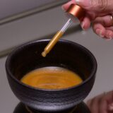 A person elegantly holds a syringe, pouring liquid into a bowl labeled 'Gold Coast Overnight Balm'.