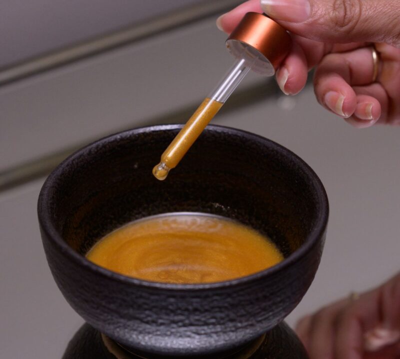 Gold Coast Overnight Balm Liquid A person elegantly holds a syringe, pouring liquid into a bowl labeled 'Gold Coast Overnight Balm'.