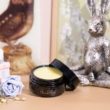 A jar of Naked Ethnobody Butter lip balm beside an open book and a bouquet of fresh flowers, exuding elegance and tranquility.